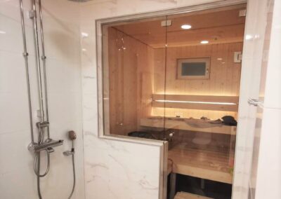 Kylpyhuoneessa valkoinen iso seinälaatta marmorikuviolla, lattiassa pieni harmaa laatta, rosterinen sadesuihku, lasiseinän läpi näkyy vaalea kuusipaneelinen sauna upotetulla kiukaalla.