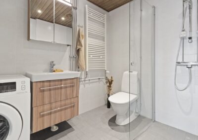 Kylpyhuone, jossa tammikuvioinen allaskaappi, valkoinen allas, valkoinen iso pyyhekuivain, vaalea iso seinälaatta, harmaa lattialaatta.